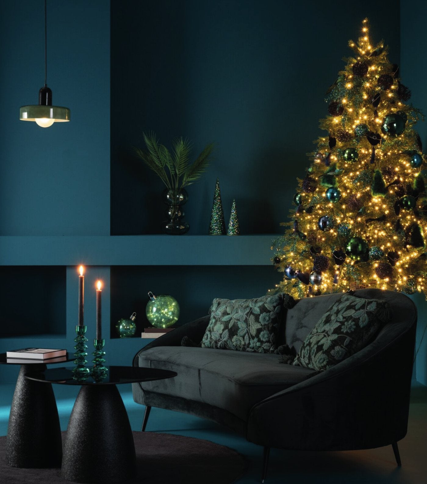 A deep teal living room dressed for Christmas with a softly glowing tree, candles and a velvet sofa